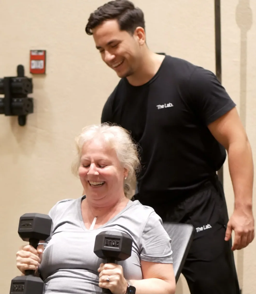 Una miembro sonriendo mientras levanta dos mancuernas, con su mentor al lado, en The Lab
